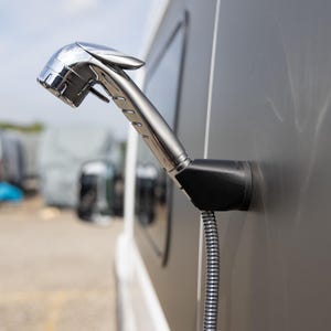 Może przedstawiać: Chromowana głowica prysznicowa przymocowana do czarnej podstawy z boku pojazdu. Głowica prysznicowa jest ustawiona pod kątem i ma żebrowany uchwyt. Elastyczny, srebrny wąż wychodzi z podstawy. W tle widać inne pojazdy.