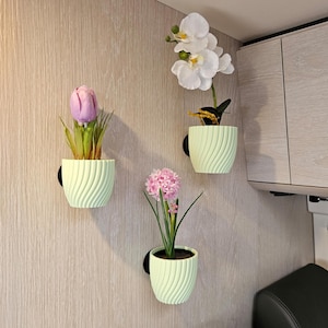 Könnte beinhalten: Drei Wandhalterungen in Hellgrün mit Spiralmuster, jede mit einer anderen künstlichen Blume: einer rosa Tulpe, einer rosa Hyazinthe und einer weißen Orchidee. Die Pflanzgefäße sind an einer hellen Wand mit Holzmaserung befestigt.