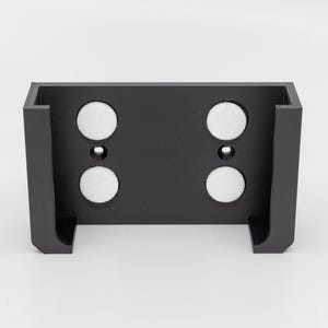 May include: A black rectangular bracket with four white circular magnets. The bracket has a U-shaped design with a flat back and two small holes. The object is likely for mounting or holding items.