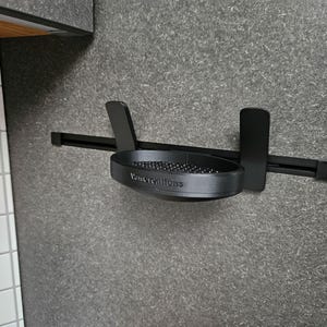 Könnte beinhalten: Ein schwarzer, an der Wand montierter Haartrocknerhalter. Der Halter hat eine runde Ablage mit kleinen schwarzen Perlen und dem Text "VanCreations". Er ist an einer strukturierten grauen Wand befestigt.