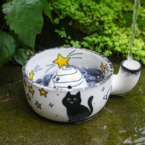 May include: A white ceramic bowl with a handle, decorated with black cat illustrations, yellow stars, and a planet. Inside the bowl sits a white, dome-shaped strainer with black paw prints, filled with blueberries and water from a stream.