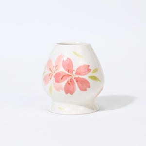 Op de afbeelding: Een kleine, crèmekleurige keramische vaas met een afgeronde body en een uitlopende voet. De vaas is versierd met handgeschilderde roze kersenbloesem bloemen en groene bladeren. De vaas is ongeveer 10 cm hoog.
