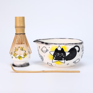 May include: A matcha tea set comprising a white ceramic bowl with a black cat design, a bamboo whisk, and a bamboo scoop. The bowl has a black rim and is decorated with stars and planets. The whisk and scoop are made of natural bamboo.