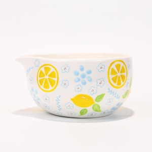 以下が含まれることがあります： 注ぎ口が付いた白いセラミック製のミキシングボウル。黄色のレモンスライス、丸ごとのレモン、青い花、緑の葉で装飾されています。ボウルは丸みを帯びた形状で、キッチンでの使用に適しています。
