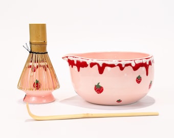 以下が含まれることがあります： ピンク色のセラミック抹茶ボウルと泡立て器のセット、竹のスクープ付き。ボウルはイチゴのデザインで、赤い滴るアクセントが付いています。泡立て器は竹製で、ピンク色のベースにイチゴのアクセントが付いています。