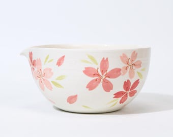 May include: A white ceramic mixing bowl with a pouring spout, adorned with hand-painted pink cherry blossom flowers and green leaves. The bowl has a wide, rounded shape, perfect for kitchen use.