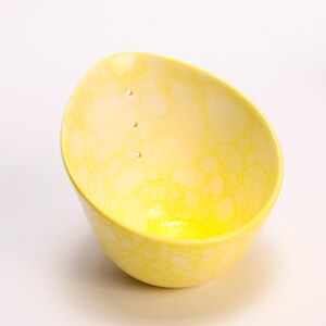 May include: A small, asymmetrical ceramic bowl featuring a bright yellow and white bubble-like pattern across its surface. The interior of the bowl includes three small, vertically aligned circular holes near the upper rim, creating a unique design.