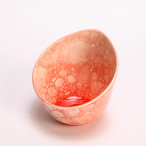May include: A small, asymmetrical ceramic bowl featuring a vibrant coral and white bubble-like pattern on the interior. The base of the bowl is a deep, solid red, and the upper rim includes three small, vertically aligned circular perforations.