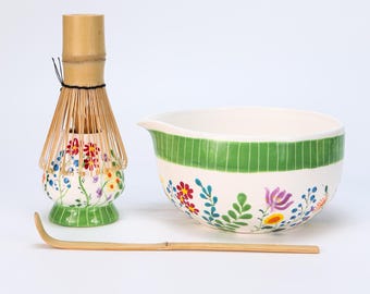 May include: A matcha tea set featuring a bamboo whisk, a ceramic bowl with a green stripe, and a bamboo scoop. The bowl and whisk holder are decorated with colorful floral patterns on a white background.