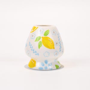 以下が含まれることがあります： 丸みを帯びた形状と小さなベースを備えた、白いセラミック抹茶泡立て器ホルダー。 黄色のレモン、緑の葉、青い花のアクセントで装飾されています。 デザインは手描きです。