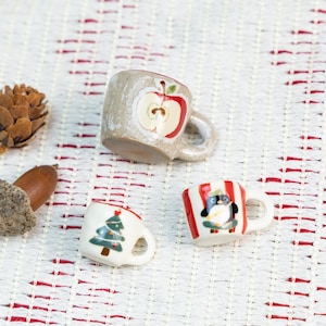 Peut inclure: Trois petites tasses en céramique avec des motifs différents. Une tasse a un motif de pomme rouge, une autre a un motif de sapin de Noël vert et la troisième a un motif rayé rouge et blanc avec un pingouin.