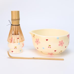 Puede incluir: Un juego de té matcha con un batidor de bambú, un cuenco de cerámica y una cuchara de bambú. El cuenco y el soporte del batidor, de color crema, están decorados con flores rosas, corazones rojos y flores azules. El batidor es de bambú.