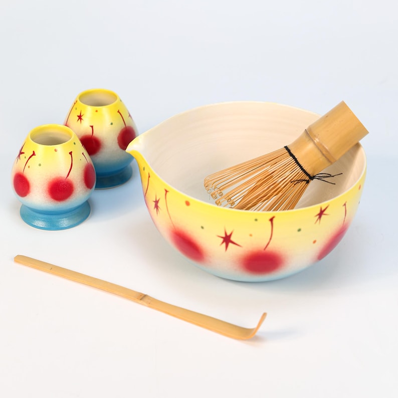 K&ouml;nnte beinhalten: Ein Matcha-Teeset mit einer gelben Schale, zwei kleinen Tassen, einem Bambusbesen und einem Bambusl&ouml;ffel. Die Schale und die Tassen haben ein Kirschdesign mit roten Kirschen und gelben Sternen. Die Tassen haben blaue B&ouml;den.