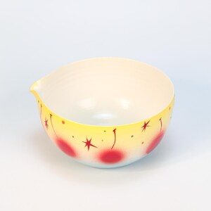 May include: A vintage-style mixing bowl with a yellow exterior, adorned with red cherries and starburst designs. The bowl has a pouring spout and a light blue base. The interior is white.