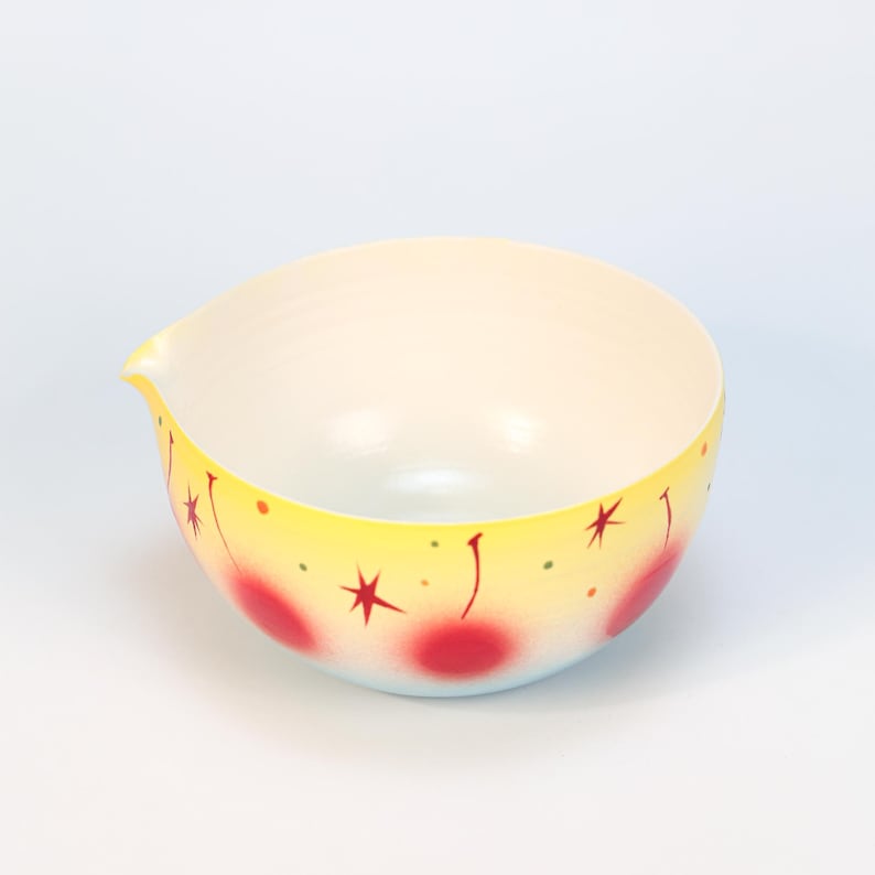 May include: A vintage-style mixing bowl with a yellow exterior, adorned with red cherries and starburst designs. The bowl has a pouring spout and a light blue base. The interior is white.