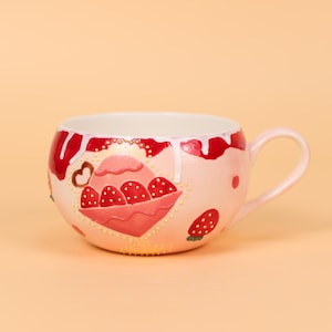 May include: A pink ceramic mug with a handle, decorated with a strawberry design. The mug features a white interior and red accents, including dripping paint and strawberry illustrations. The mug is on a peach-colored background.