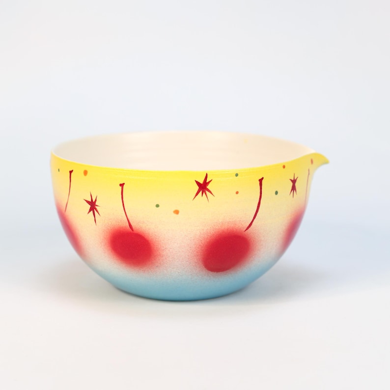 May include: A vintage mixing bowl with a yellow, red, and blue gradient design. The bowl features red cherry designs and starburst accents. It has a pouring spout and a white interior.