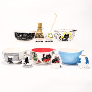 以下が含まれることがあります： 気まぐれな猫のデザインが施されたセラミックボウルとアクセサリーのコレクション。ボウルには、白黒のパターン、猫のイラスト、スイカのスライスが描かれています。竹製の泡立て器と青いボウルが含まれています。