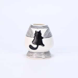 May include: A white ceramic matcha bowl with a black cat design. The bowl features a black cat silhouette on the front, with black striped patterns around the top and bottom. The bowl has a rounded shape and a small base.