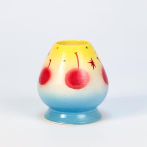 K&ouml;nnte beinhalten: Eine handbemalte Keramikvase mit gelbem Oberteil, das in Wei&szlig; und dann in Blau am Boden &uuml;bergeht. Die Vase hat rote Kirschmotive und einen Stern. Die Vase ist etwa 10 cm hoch.