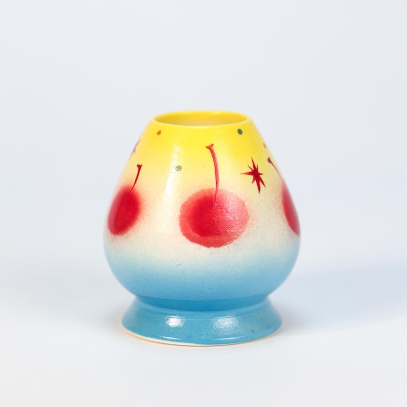 K&ouml;nnte beinhalten: Eine handbemalte Keramikvase mit gelbem Oberteil, das in Wei&szlig; und dann in Blau am Boden &uuml;bergeht. Die Vase hat rote Kirschmotive und einen Stern. Die Vase ist etwa 10 cm hoch.