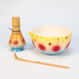 K&ouml;nnte beinhalten: Ein Matcha-Teeset mit einem Bambusbesen, einem Bambusl&ouml;ffel und einer Keramikschale. Die Schale ist gelb mit roten Kirschmotiven und einem blauen Sockel. Der Schneebesen hat einen hellbraunen Griff und ein gelbes Schneebesenende.