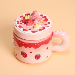 May include: A pink ceramic mug with a lid, decorated to resemble a strawberry cake. The mug features a handle and is adorned with painted strawberries, sprinkles, and frosting details. The lid is topped with strawberry and cream decorations.