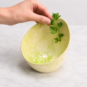 May include: A light yellow ceramic bowl with a vibrant green bubble-like pattern on the interior surface. The bowl features a small circular opening near the rim, through which a sprig of fresh green parsley is inserted and held upright.