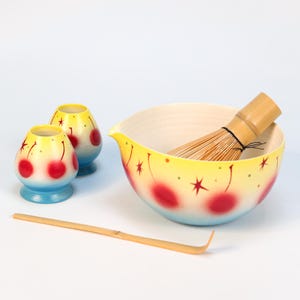 K&ouml;nnte beinhalten: Ein Matcha-Teeset mit einer gelben Schale und zwei kleinen Tassen, alle mit roten Kirschen und Sternen verziert. Ein Bambusbesen und -l&ouml;ffel sind ebenfalls enthalten. Die Schale hat einen Durchmesser von etwa 15 cm.