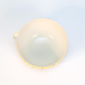 May include: A ceramic bowl with a light yellow exterior and a white interior. The bowl has a small spout on one side and a decorative yellow rim with red and purple accents. The bowl is likely used for serving food.