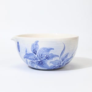 May include: White ceramic bowl with a spout, decorated with blue floral designs. The bowl features a hand-painted lily pattern, with detailed petals and stamen. The bowl is ideal for serving or display.