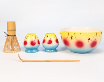 May include: A collection of matcha tea ceremony tools. Includes a bamboo whisk, two small ceramic containers, a bamboo scoop, and a large bowl. The ceramic pieces have a blue, yellow, and red cherry design.