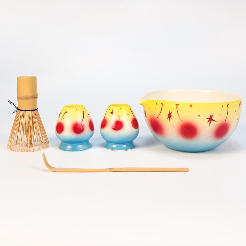 K&ouml;nnte beinhalten: Eine Sammlung von Matcha-Tee-Zeremonie-Werkzeugen. Enth&auml;lt einen Bambusbesen, zwei kleine Keramikbeh&auml;lter, einen Bambusl&ouml;ffel und eine gro&szlig;e Sch&uuml;ssel. Die Keramikteile haben ein blaues, gelbes und rotes Kirschdesign.