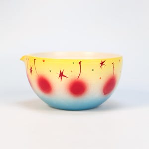 Może przedstawiać: Miska do mieszania w stylu vintage z żółtą górą, która przechodzi w niebieski na dole. Miska ma czerwone wiśnie i wzory gwiazd. Ma dzióbek do nalewania z boku.
