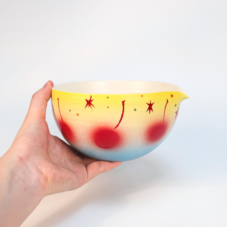 May include: A vintage mixing bowl with a yellow, red, and blue gradient design. The bowl features red cherry and star illustrations. The bowl has a pouring spout and is held by a hand.
