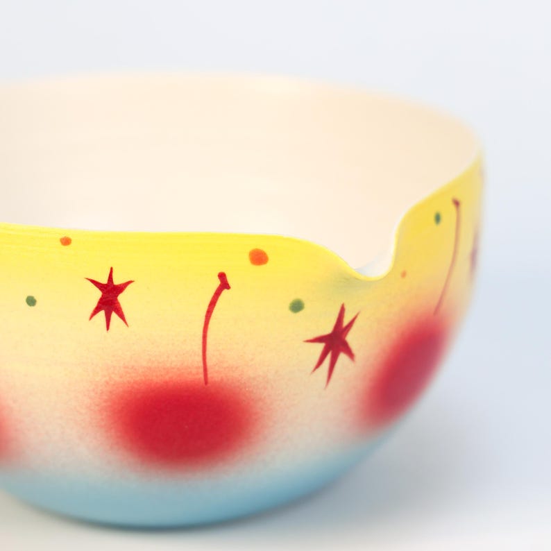 May include: A decorative bowl with a yellow rim and a light blue base. The bowl is decorated with red star and dot designs. The interior of the bowl is white.