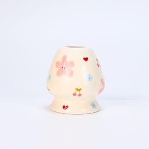 May include: A cream-colored ceramic vase with a rounded body and a flared base. The vase is decorated with pink flowers, red hearts, blue flowers, and yellow cherries. The vase has a smooth, glossy finish.