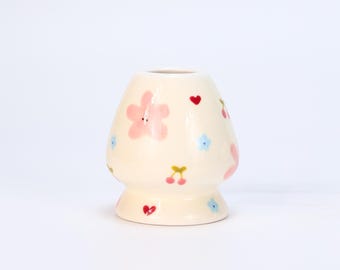 May include: A cream-colored ceramic vase with a rounded body and a flared base. The vase is decorated with pink flowers, red hearts, blue flowers, and yellow cherries. The vase has a smooth, glossy finish.