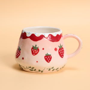 May include: A ceramic mug with a pink base, adorned with red strawberries and green leaves. The rim features a red and white design. The handle is pink, and the mug has a capacity of approximately 12 ounces.