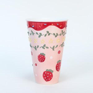 May include: A tall, pink ceramic tumbler with a white interior. The tumbler is decorated with red strawberries, yellow flowers, and green vines. The rim has a red wavy design with blue and white sprinkles.