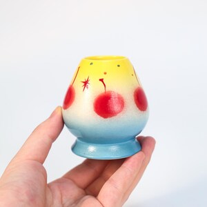 K&ouml;nnte beinhalten: Kleine, handbemalte Keramikvase mit rundem K&ouml;rper und ausgestelltem Fu&szlig;. Die Vase hat einen Farbverlauf von Gelb nach Blau mit roten Kirschmotiven. Die Vase wird in einer Hand vor wei&szlig;em Hintergrund gehalten.