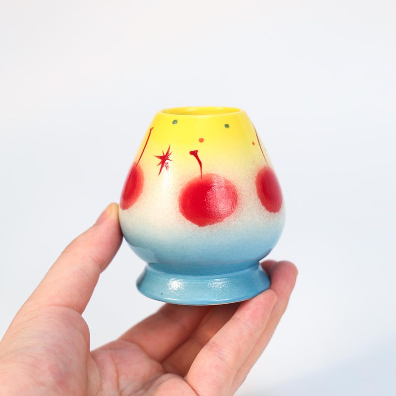 K&ouml;nnte beinhalten: Kleine, handbemalte Keramikvase mit rundem K&ouml;rper und ausgestelltem Fu&szlig;. Die Vase hat einen Farbverlauf von Gelb nach Blau mit roten Kirschmotiven. Die Vase wird in einer Hand vor wei&szlig;em Hintergrund gehalten.