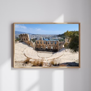 May include: A framed photograph of an ancient amphitheater with tiered seating and a stage. The structure is made of light-colored stone, with a backdrop of a city and a clear blue sky. The image is in a wooden frame.