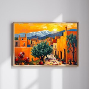 May include: A framed art print showing a colourful street scene. The artwork features orange buildings, green trees, and a patterned pathway. A bright yellow sun sets behind snow-covered mountains. Decorative red pots are scattered throughout the scene.