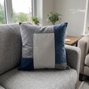 May include: A square cushion with a patchwork design in shades of grey and navy blue. The central panel is light grey, surrounded by darker grey and navy blue velvet sections. The cushion is on a grey sofa.
