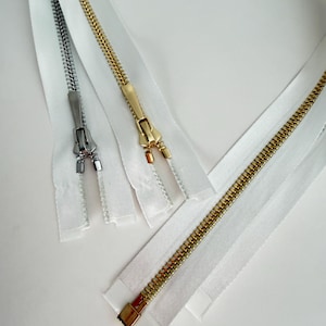 May include: Three white fabric zippers with metal teeth and sliders. One zipper features silver-toned hardware, while the other two zippers feature gold-toned hardware. Each zipper has a long white fabric tape and a metal pull tab for operation.