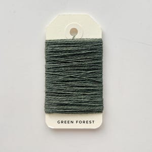 May include: A spool of dark green thread labeled "GREEN FOREST" on a cream-colored tag. The thread is tightly wound, showcasing a textured surface. The tag has a hole at the top for hanging. Ideal for crafting and sewing projects.