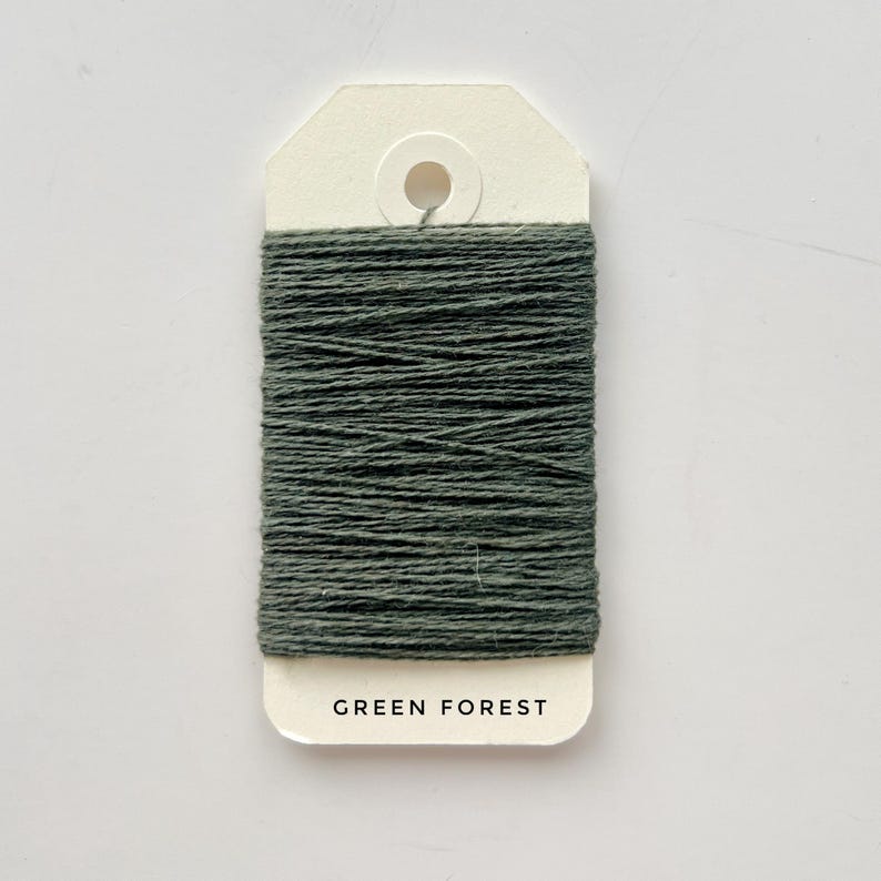 May include: A spool of dark green thread labeled "GREEN FOREST" on a cream-colored tag. The thread is tightly wound, showcasing a textured surface. The tag has a hole at the top for hanging. Ideal for crafting and sewing projects.