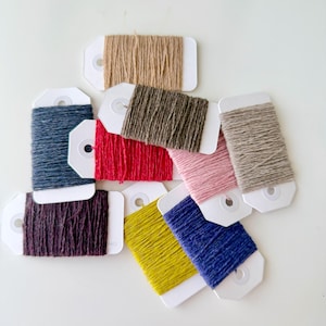 Peut inclure: Plusieurs bobines de fil colorées sur des étiquettes blanches. Les bobines présentent une variété de couleurs, notamment marron, rouge, rose, jaune, bleu et violet. Le fil est étroitement enroulé sur les étiquettes, créant un aspect texturé. Les étiquettes sont blanches avec un petit trou en haut.