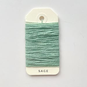 May include: A spool of sage green thread is wrapped around a cream-colored tag. The word "SAGE" is printed in black letters at the bottom of the tag. The thread is tightly wound, creating a textured appearance.